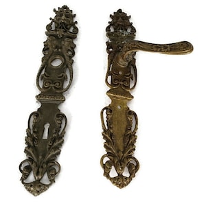 Può includere: Due maniglie per porte in ottone antico con intricati disegni floreali e a volute. Le maniglie sono ornate e presentano un buco della serratura e una leva.