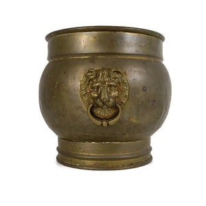 May include: A large, round, brass planter with a lion's head design on the front. The planter has a slightly distressed finish and a wide, flat base.