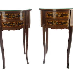 Puede incluir: Dos mesas auxiliares de madera a juego con tapas curvas y dos cajones. Las mesas tienen intrincados diseños de marquetería en tonos marrones, dorados y negros. Cada mesa tiene tres patas.