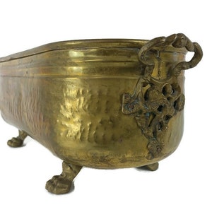 May include: A brass planter with an ornate design and three feet. The planter has a hammered finish and a decorative handle.