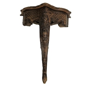 May include: Ornate, antique-style wall shelf in a warm, neutral tone. The shelf features intricate carvings and a single, elongated leg. The design includes diamond and floral patterns, creating a classic, vintage aesthetic.
