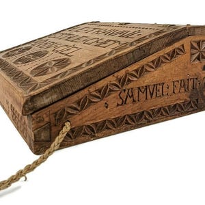 May include: A wooden box with a hinged lid and a carved inscription that reads "Le Travail Est Le Chevet Du Bon Ciel" and "Samvel Fait". The box is decorated with a carved floral pattern and has a rope handle.