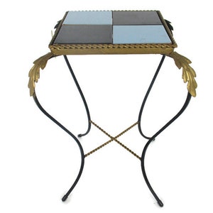 Può includere: Un piccolo tavolino quadrato con un piano in piastrelle nere e blu. Il tavolo ha un bordo dorato e dettagli in foglia d'oro sulle gambe. Le gambe sono in metallo nero e hanno un design intrecciato.