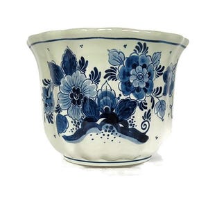 May include: A white ceramic planter with a scalloped rim, decorated with hand-painted blue floral designs. The flowers and leaves are arranged in a repeating pattern around the exterior. A decorative ribbon design is also present.
