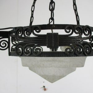 May include: Black wrought iron chandelier with a frosted glass shade. The fixture features decorative scrollwork and is suspended by chains. The shade has a tiered, geometric design, and the light bulbs are visible.