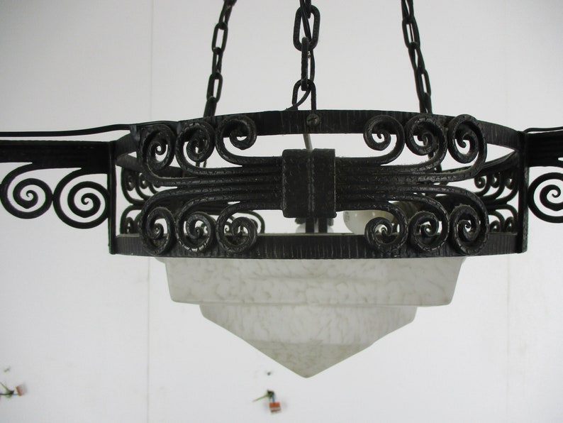 May include: Black wrought iron chandelier with a frosted glass shade. The fixture features decorative scrollwork and is suspended by chains. The shade has a tiered, geometric design, and the light bulbs are visible.