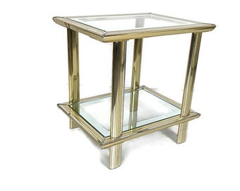 May include: A vintage side table with a brass-colored metal frame and two glass shelves. The table has a square shape and is supported by four cylindrical legs. The glass shelves are clear, and the metal frame has a polished finish.
