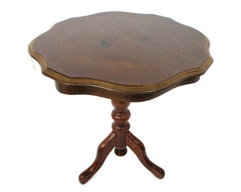 Op de afbeelding: Een donkerbruine bijzettafel in antieke stijl met een geschulpte rand. De tafel heeft een rond blad met een decoratieve rand en een voetstuk met drie gebogen poten. De tafel is gemaakt van hout en heeft een glanzende afwerking.