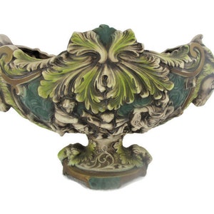 May include: A large, ornate, green and gold ceramic bowl with a sculpted design of leaves and figures. The bowl is on a pedestal base.