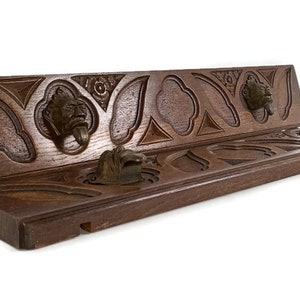 Pode incluir: Um cabide de parede de madeira com entalhes intrincados e quatro ganchos de metal. O cabide é tingido de uma cor marrom escura e tem um visual rústico e antigo.