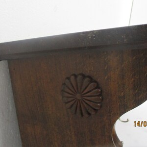 May include: A dark brown wooden shelf with a decorative floral carving. The shelf has a curved lower edge and a metal loop on the side for hanging. The wood grain is visible, and the shelf appears to be vintage.
