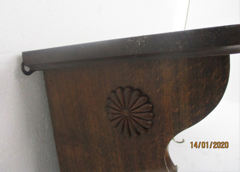 May include: A dark brown wooden shelf with a decorative floral carving. The shelf has a curved lower edge and a metal loop on the side for hanging. The wood grain is visible, and the shelf appears to be vintage.