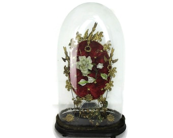 May include: A glass dome with a velvet heart-shaped base, decorated with gold-coloured metal birds and leaves. Inside the dome, a white rose and other flowers are displayed on a red velvet cushion.