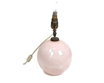 May include: A small, round, pink ceramic table lamp with a brass-coloured base and bulb socket. The lamp has a white cord and a simple on/off switch. The lamp is a sphere shape.