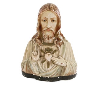 Puede incluir: Un busto de yeso blanco de Jesucristo con un corazón rojo y espinas en el pecho. Su cabello y barba son marrones y lleva una túnica blanca.
