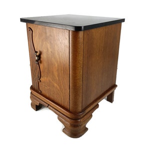 Puede incluir: Una pequeña mesita de noche de madera con una tapa negra y un solo cajón. El cajón tiene una manija de metal decorativa y la mesita de noche tiene patas talladas.