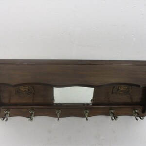 May include: A dark brown wooden wall shelf with a mirror and six metal hooks. The shelf has decorative carvings and a sloped top. A practical storage solution for hallways or entryways, measuring approximately 100cm in length.
