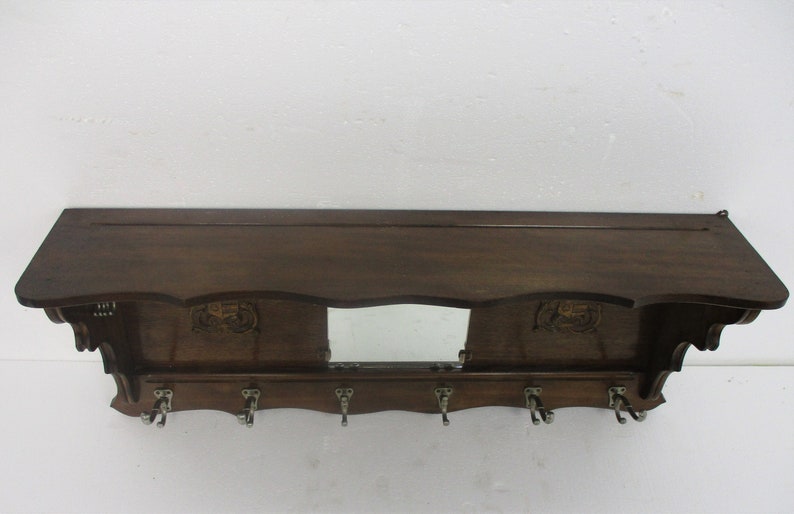 May include: A dark brown wooden wall shelf with a mirror and six metal hooks. The shelf has decorative carvings and a sloped top. A practical storage solution for hallways or entryways, measuring approximately 100cm in length.