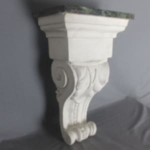 May include: White, ornate wall shelf with a green marble top. The shelf has a decorative, scrolled corbel design and is made of a solid, possibly plaster, material. The shelf is designed to be mounted on a wall.