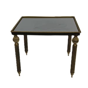 Puede incluir: Una mesa auxiliar cuadrada con tapa negra y ribete dorado. La mesa tiene cuatro patas con un diseño a rayas doradas y negras y detalles dorados decorativos. La mesa probablemente está hecha de madera y metal.