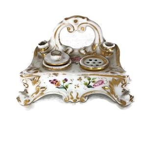 May include: An antique white porcelain inkwell with gold accents and floral details. The inkwell features two ink holders, a central pen rest, and a decorative arched handle. The base is adorned with painted flowers.