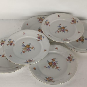 May include: Six white ceramic plates with scalloped edges and gold rims. Each plate features a central floral bouquet design in shades of orange, blue, yellow, and purple. A classic tableware set.