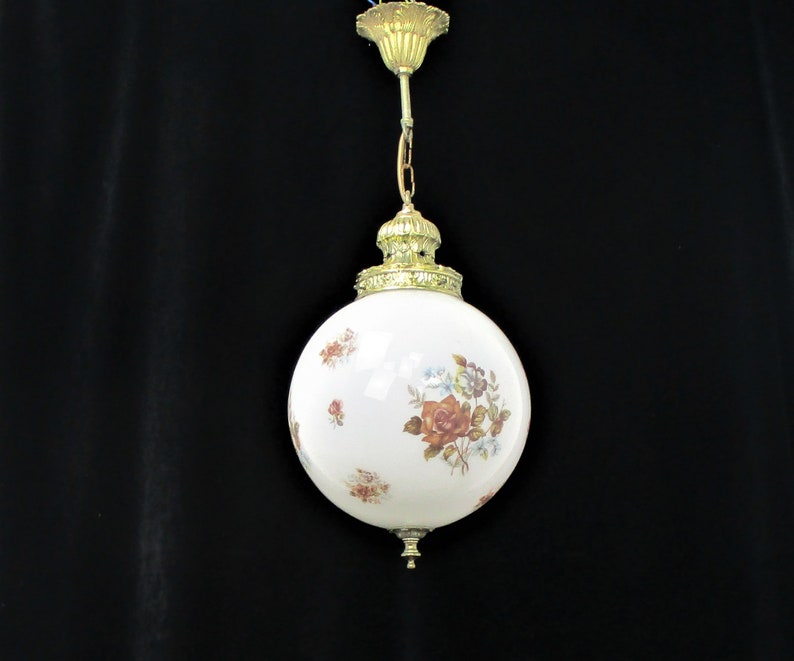 May include: A vintage globe pendant light with a white glass shade adorned with floral patterns in shades of red, brown, and blue. The light fixture features brass-colored metal accents and a decorative top.