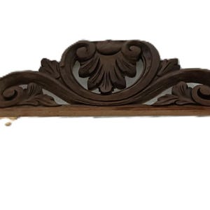 Può includere: Un elemento architettonico in legno marrone scuro, ornato con un motivo floreale centrale e dettagli a volute. Il pezzo ha una base piatta e un piccolo supporto rettangolare che si estende dal basso.