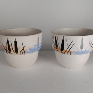 Peut inclure: Deux pots en céramique blanche avec un motif abstrait bleu, orange et noir. Le motif représente des roseaux ou des quenouilles stylisés.
