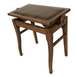 Puede incluir: Un taburete de piano de madera vintage con un asiento rectangular de cuero marrón. El taburete tiene patas curvas y un mecanismo para ajustar la altura del asiento. La madera tiene un acabado cálido y natural.
