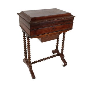 Può includere: Un'antica tavola da cucito marrone scuro con due cassetti e un ripiano inferiore. Il tavolo ha gambe tornite e un bordo perlato attorno alla parte superiore. Il tavolo è alto circa 76 cm.