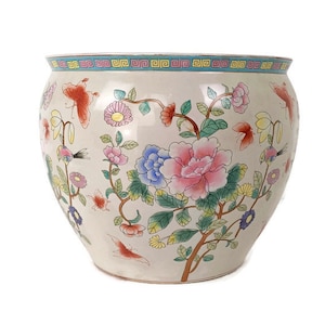 Könnte beinhalten: Ein weißer Keramiktopf mit Blumenmuster. Der Topf zeigt rosa, blaue, gelbe und grüne Blumen mit Schmetterlingen und Vögeln. Der Rand des Topfes hat ein blaues und gelbes geometrisches Muster.