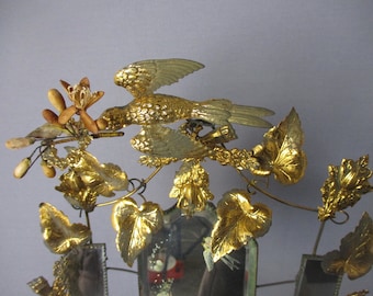 May include: A gold-toned metal bird with outstretched wings perched on a branch with gold-toned leaves. The bird is holding a small, dried flower in its beak. The branch is attached to a mirror with a gold-toned frame.