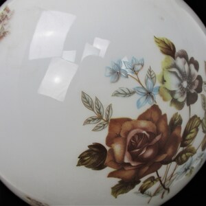May include: A white, round glass object with floral designs. The flowers are in shades of brown, blue, and yellow, with green leaves. The object has a glossy finish and is set against a black background.