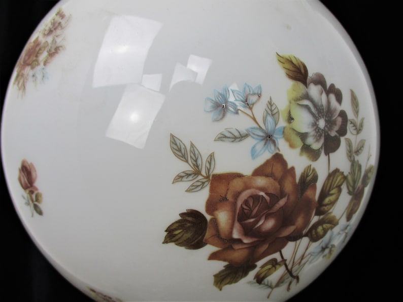 May include: A white, round glass object with floral designs. The flowers are in shades of brown, blue, and yellow, with green leaves. The object has a glossy finish and is set against a black background.