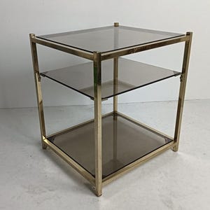 May include: A three-tiered side table with a gold-colored metal frame and three smoked glass shelves. The table has a square shape and is designed for display or storage. The table measures approximately 20 inches in height, 20 inches in width, and 20 inches in depth.