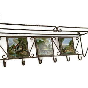 Könnte beinhalten: Ein Vintage-Wandgarderobe aus Metall mit einem Regal und drei Landschaftsflieseneinsätzen. Der dunkelbraune Ständer verfügt über dekorative Schnörkel und vier Haken zum Aufhängen von Gegenständen. Die Fliesen zeigen ruhige Szenen im Freien.