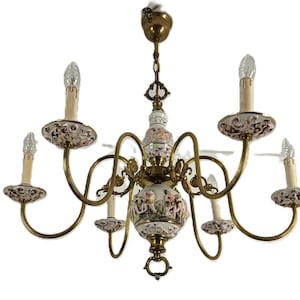 May include: An antique chandelier with six candle-style lights. The fixture has a brass finish with decorative porcelain elements featuring floral and figural designs. The candle holders have a cream color and the chandelier is suspended from a chain.