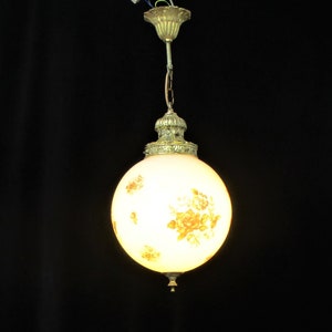May include: A vintage-style pendant light with a spherical, frosted glass shade. The shade is adorned with a floral pattern in shades of yellow and orange. The fixture has ornate brass-colored metal detailing and a chain for hanging.