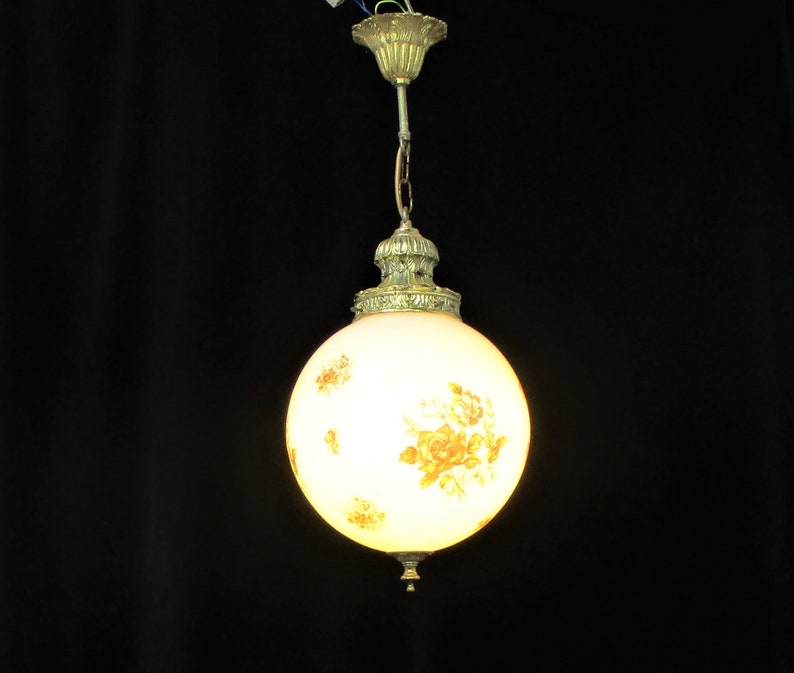May include: A vintage-style pendant light with a spherical, frosted glass shade. The shade is adorned with a floral pattern in shades of yellow and orange. The fixture has ornate brass-colored metal detailing and a chain for hanging.
