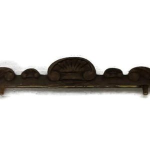 May include: A dark brown, ornate furniture hardware piece with a symmetrical design. The piece features a central, fan-shaped element flanked by scrolling details and small, round accents. Two small pegs extend from the back.