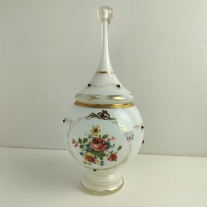 Peut inclure: Pot en verre blanc avec un motif floral et des accents dorés. Le pot a un couvercle avec un haut pointu et haut.