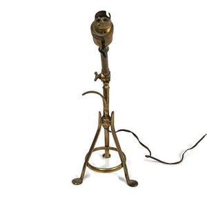 May include: A vintage brass desk lamp with an adjustable arm and a tripod base. The lamp has a classic design with a bulb socket at the top and a black power cord. The lamp is made of brass and has a warm, antique finish.