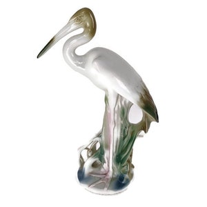 Puede incluir: Una figurita de porcelana de una garza blanca con detalles marrones y verdes de pie sobre una base con detalles verdes y rosas.