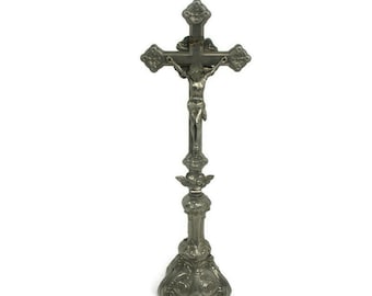 May include: A silver-colored crucifix with a detailed sculpture of Jesus Christ. The cross has ornate detailing on the arms and base, and stands on a decorative pedestal. The religious item is made of metal.