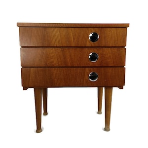 Peut inclure: Une table de chevet en bois à trois tiroirs avec une finition brun clair et des poignées de tiroir en chrome. La table de chevet a quatre pieds.