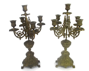 May include: Two antique brass candelabras with ornate designs. Each candelabra has multiple arms for candles, with detailed floral and scrollwork patterns. The bases are also intricately designed, showcasing a vintage aesthetic.