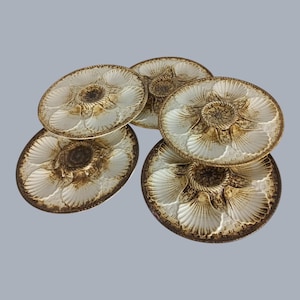 May include: A collection of five white and brown oyster plates. Each plate features a central well and radiating shell-shaped compartments. The plates have a textured, detailed design with a brown rim and center.