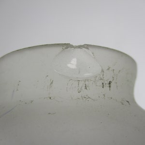 May include: A close-up of a frosted glass bottle with a damaged rim. The bottle has a rounded shape with a narrow neck and a wider base. The glass has a slightly textured surface and appears to be translucent.
