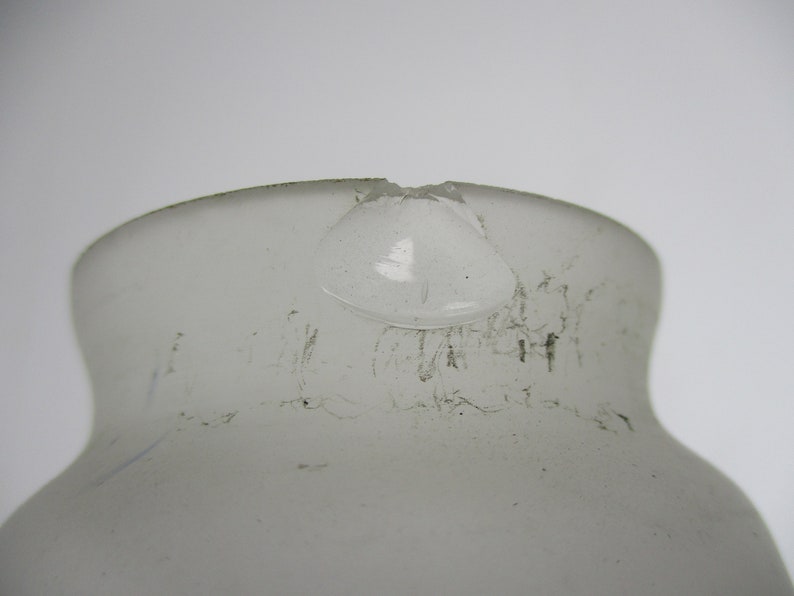 May include: A close-up of a frosted glass bottle with a damaged rim. The bottle has a rounded shape with a narrow neck and a wider base. The glass has a slightly textured surface and appears to be translucent.